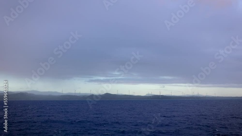 Wallpaper Mural Ocean coastline with wind power turbines spread on the shore during stormy, cloudy day with blue cast as seen from a ship sailing along them Torontodigital.ca