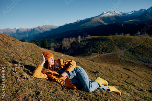 Woman lying on a hill on the grass rest smile with teeth looking at the mountains in the snow in winter in a yellow raincoat and jeans happy sunset trip on a hike, freedom lifestyle 