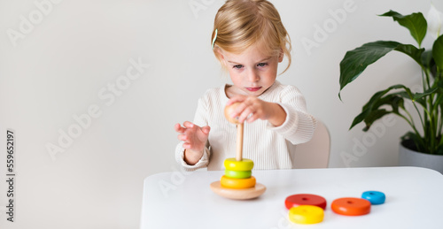 Adorable blonde toddler, kid, child, girl two years old playing indoor with eco wooden coloured toy pyramidion on white, gray background. Copy space.Early preschool development, kindergarten game. photo