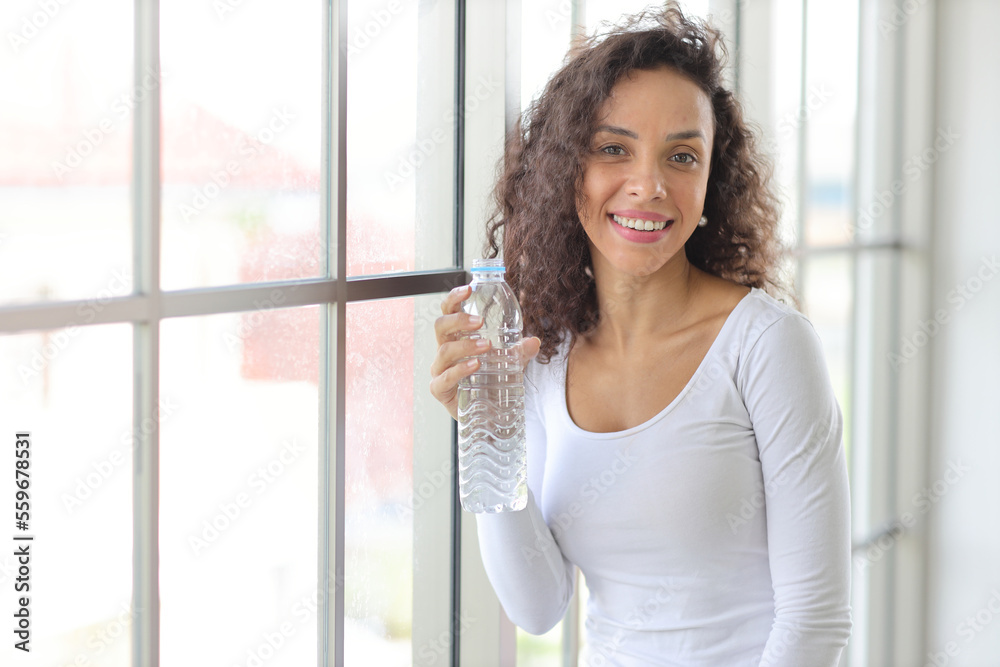 Charming African American holding and drinking bottle of water drink clean clear mineral aqua for healthy and body refreshment. Happy female standing in living room at home Charming African American holding and drinking bottle of water drink clean clear mineral aqua for healthy and body refreshment. Happy female standing in living room at home