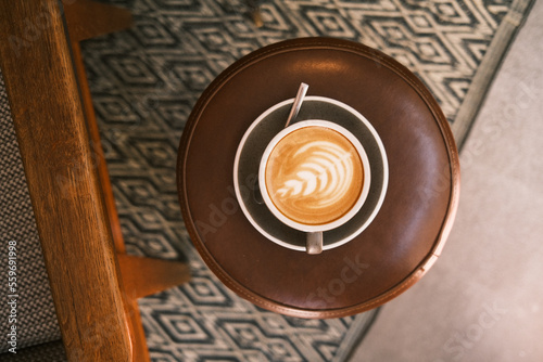 Café de barista con dibujo latte art hecho a mano y servido en taza y plato vintage sobre taburete de cuero y alfombra con patrón gométrico.