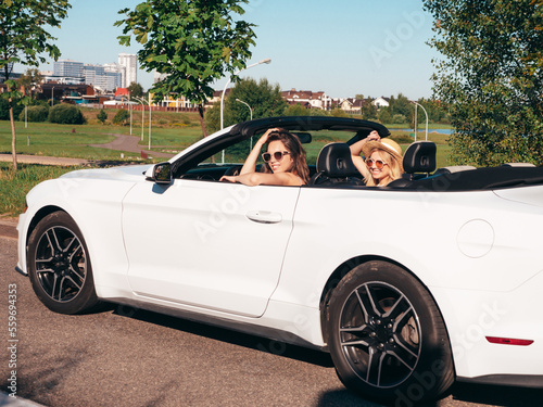 Portrait of two young beautiful and smiling hipster female in convertible car. Sexy carefree women driving cabriolet. Positive models riding and having fun in sunglasses outdoors. Enjoying summer days © halayalex