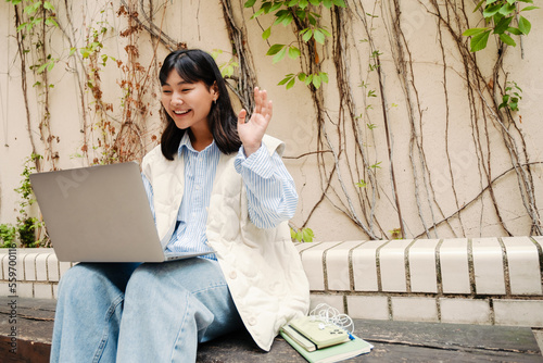 Cheerful asian girl making video call on laptop while sitting outdoors on the bench © Drobot Dean