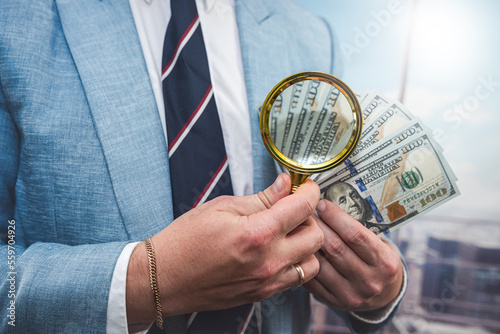 businessman wear blue suit looking with magnifying glass on dollar bills in office
