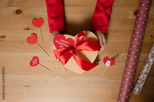 Heart-shaped box decorated with red satin ribbon in children's hands, Happy Valentine's Day card