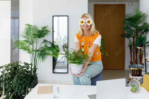 Happy woman wearing flower sunglasses sitting on chair with plant at home
