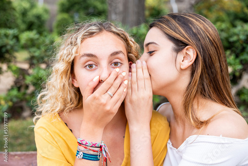 Woman whispering into shocked friend's ear in park photo