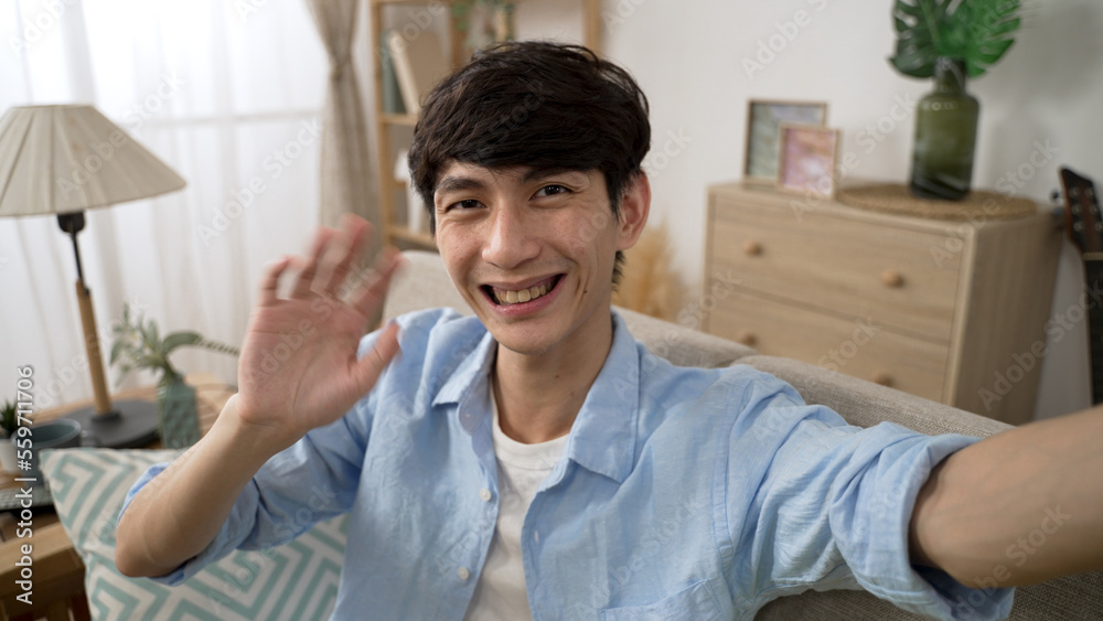 smiling asian Chinese guy facing mobile phone cam is nodding head and waving goodbye to his friend while having a video chat in the living room at home smiling asian Chinese guy facing mobile phone cam is nodding head and waving goodbye to his friend while having a video chat in the living room at home