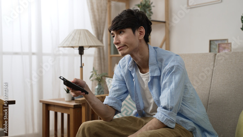 impatient asian Japanese man shaking head with dissatisfied look on face while channel surfing with a tv controller on the couch in the living room at home
