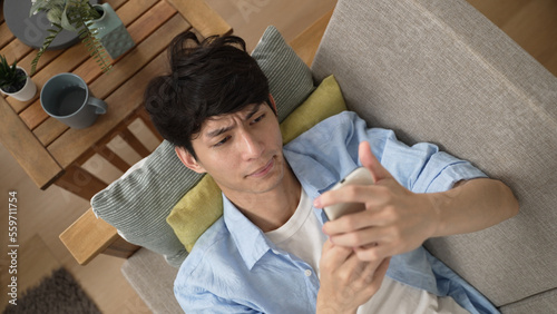 top angle view of a sleepy Korean guy dropping his cell phone while lying on the couch and browsing social media feeds in the living room at home.