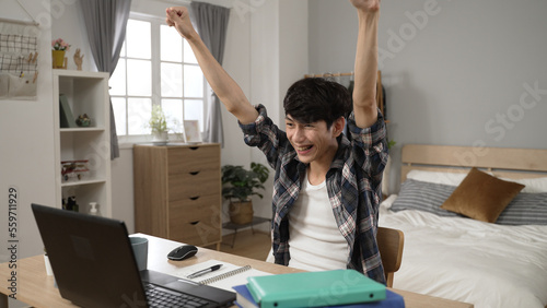 Fotografija slow motion excited asian Japanese adult student celebrating with lifting clenched fists after checking the college acceptance email on computer in his bedroom at home
