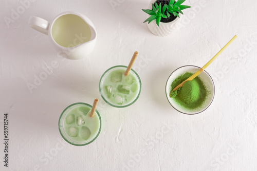 two glasses with green iced tea matcha latte, a bowl of matcha powder, a milk jug with milk. top view. cooking white background. wellness drink.