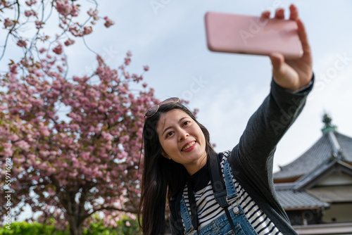 happy asian Korean girl tourist having fun chatting with friend on phone via video call with beautiful cherry blossom at background as she visits Kofuku-ji temple in nara japan
