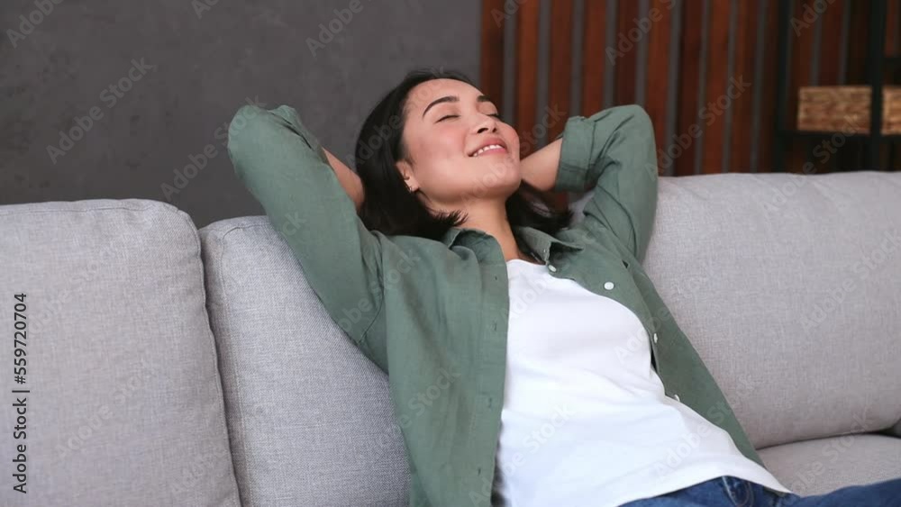 Young woman of Asian ethnicity wear casual clothes hold hands behind neck take nap with closed eyes sit on sofa stay at home hotel flat rest relax spend free spare time in living room indoor grey wall Young woman of Asian ethnicity wear casual clothes hold hands behind neck take nap with closed eyes sit on sofa stay at home hotel flat rest relax spend free spare time in living room indoor grey wall