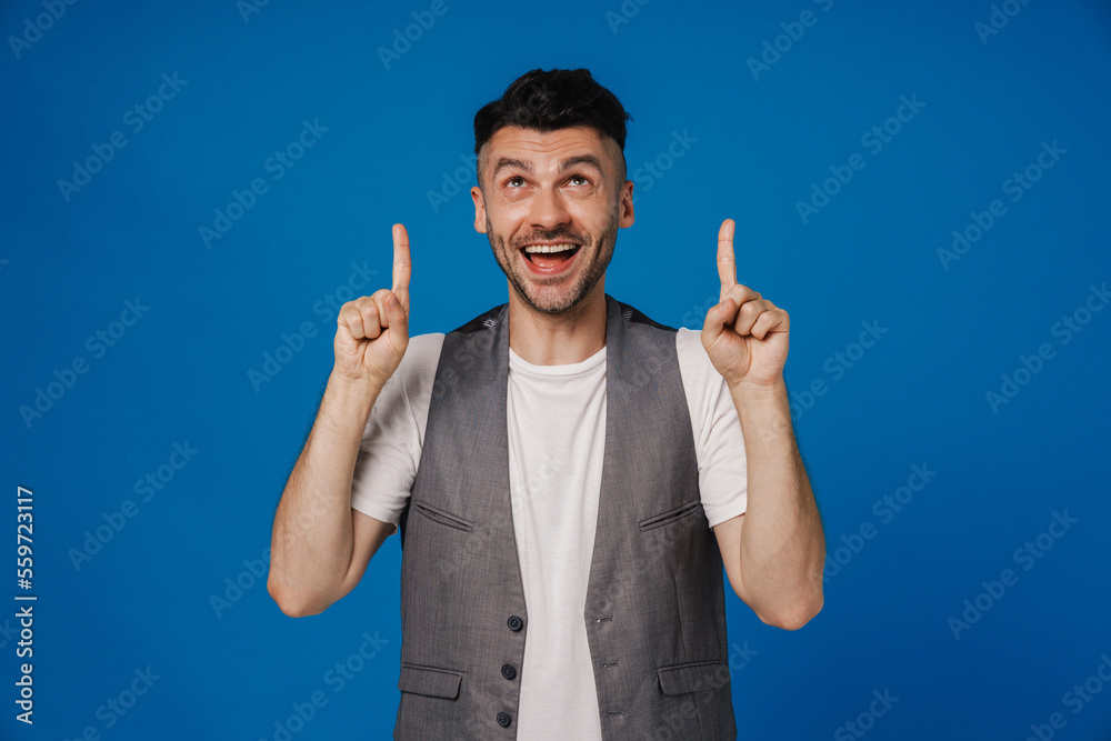 Excited man pointing fingers up at copy space isolated over blue studio wall Excited man pointing fingers up at copy space isolated over blue studio wall