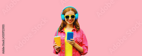 Portrait of stylish modern happy smiling young woman in headphones listening to music with smartphone wearing jacket on vivid pink background