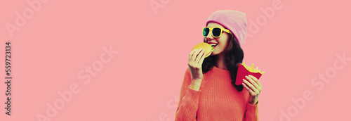 Portrait of stylish happy smiling young woman with burger and french fries fast food wearing hat on pink background © rohappy