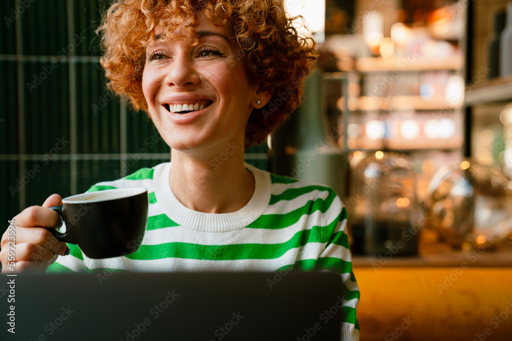 Fototapeta premium Mid white woman with curly hair smiling and drinking coffee in cafe