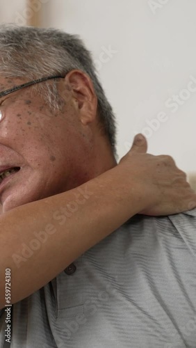 Vertical Screen: Senior asian man with  white hair sitting on couch feeling pain and massage his shoulder at home. illness old male resting on sofa with hurting neck.