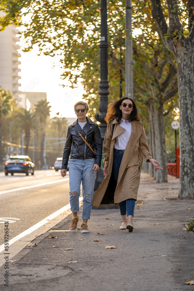 Two adults, 35s years old women with sunglasses and casual clothing, walk along the street. Urban modern lifestyle scene of lesbian and straight friends in Barcelona city.
