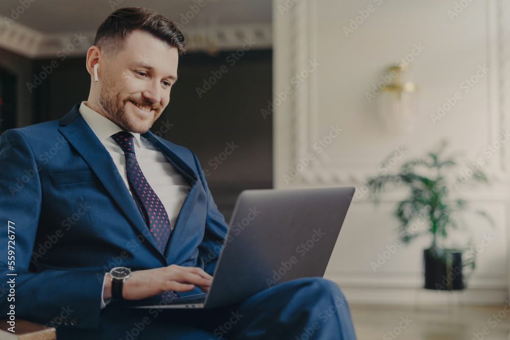 Cheerful unshaven prosperous male entrepreneur works on laptop computer works remotely has video conference dressed in formal suit with tie poses at cozy interior calls partner via online chat Cheerful unshaven prosperous male entrepreneur works on laptop computer works remotely has video conference dressed in formal suit with tie poses at cozy interior calls partner via online chat