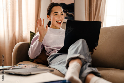 Excited smiling asian woman making video call via laptop and waving hand while sitting on couch