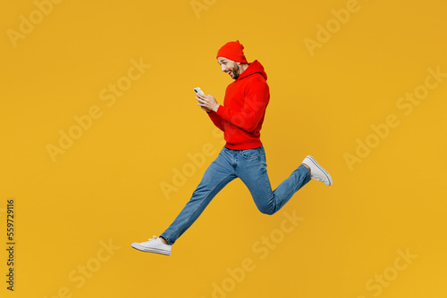 Full body side view young caucasian man wear red hoody hat jump high hold in hand use mobile cell phone chat online isolated on plain yellow color background studio portrait. People lifestyle concept.
