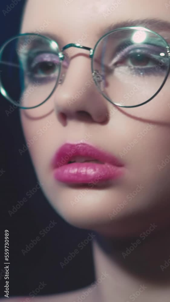 Vertical video. 90s fashion. Eyewear model. 2000s beauty look. Teen girl face with artistic makeup wearing round frame glasses in purple blue color neon light on dark. Vertical video. 90s fashion. Eyewear model. 2000s beauty look. Teen girl face with artistic makeup wearing round frame glasses in purple blue color neon light on dark.