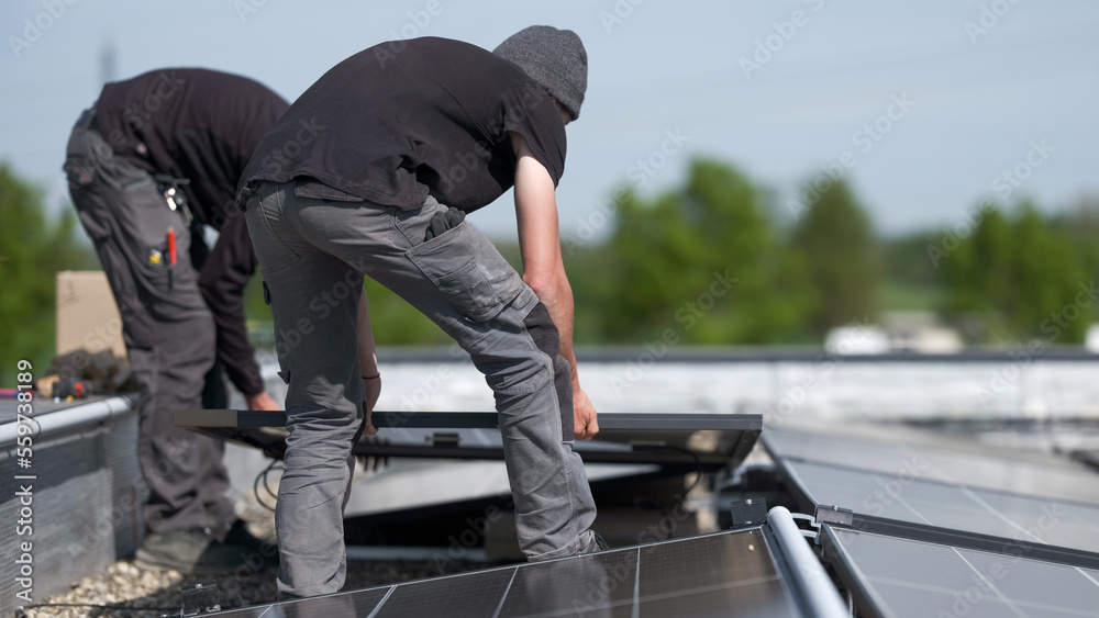 Male team engineers installing stand-alone solar photovoltaic panel system. Electricians mounting blue solar module on roof of company roof. Alternative energy concept
