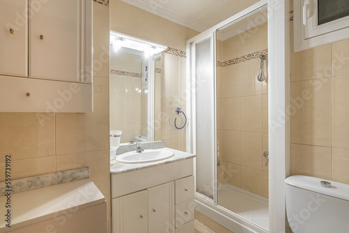 Bathroom is bright by lamps above small mirror with light beige tile walls. Open shower cubicle in white plastic with frosted glass sliding door. Dressing table with many drawers and white sink.