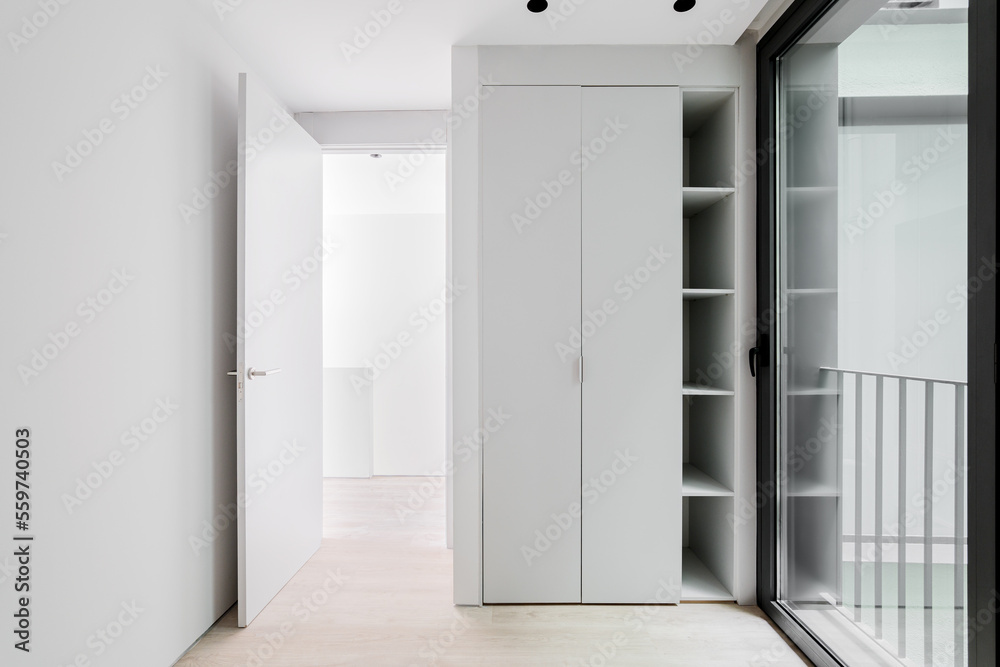 Small entrance hall for meeting guests with large glass front door in black metal frame, which reflects part of railing on street. White wooden wardrobe with closed doors and shelves for small items. Small entrance hall for meeting guests with large glass front door in black metal frame, which reflects part of railing on street. White wooden wardrobe with closed doors and shelves for small items.