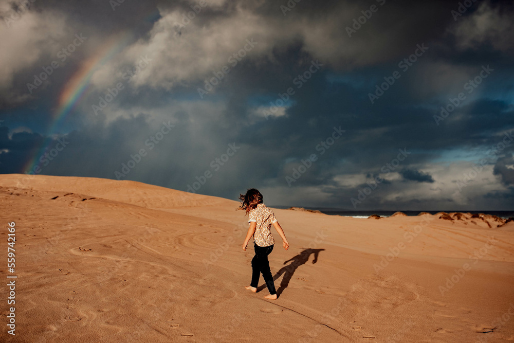 Boy walking on sand dune with rainbow in sky Boy walking on sand dune with rainbow in sky