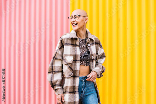 Happy woman with shaved head wearing plaid jacket standing in front of wall photo
