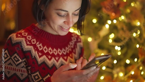 closeup cute smiling woman in red festive sweater on background of lights on christmas tree using phone, messaging, reading and writing congratulations, looking social networks. holyday mood