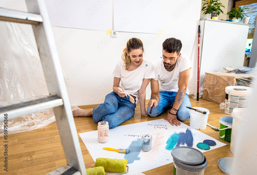 Young couple sitting on the floor choosing color for painting the wall in their home.