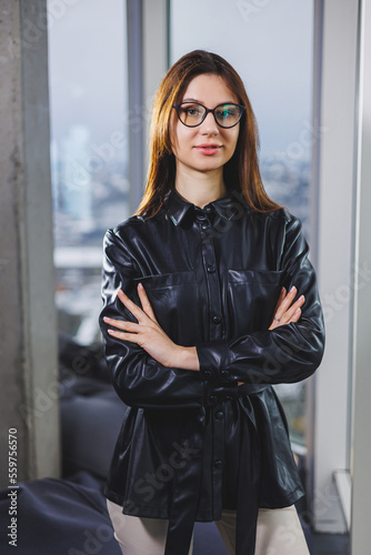 Portrait of a young slender woman in glasses and a black leather shirt. Modern woman on the background of the window in the office with a large window