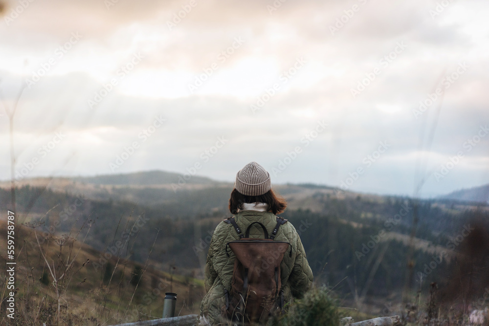 View from the back of hiker girl wearing backpack and beanie hat sitting on edge of cliff and enjoying landscape, sunset, mountains and clouds.