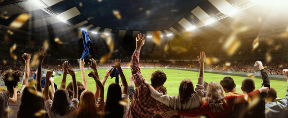 Back view of football, soccer fans cheering their team, holding flag at crowded stadium at evening time. Concept of sport, hobby, leisure time, football