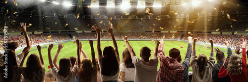 Back view of football, soccer fans emotionally cheering their team at crowded stadium at evening time. Concept of sport, hobby, leisure time, football