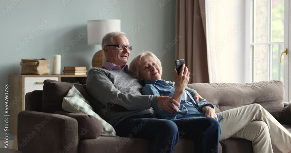 Relaxed mature wife and husband resting on couch at home look at smartphone screen, choose goods through retail website services, use mobile app, older generation and modern wireless technology usage