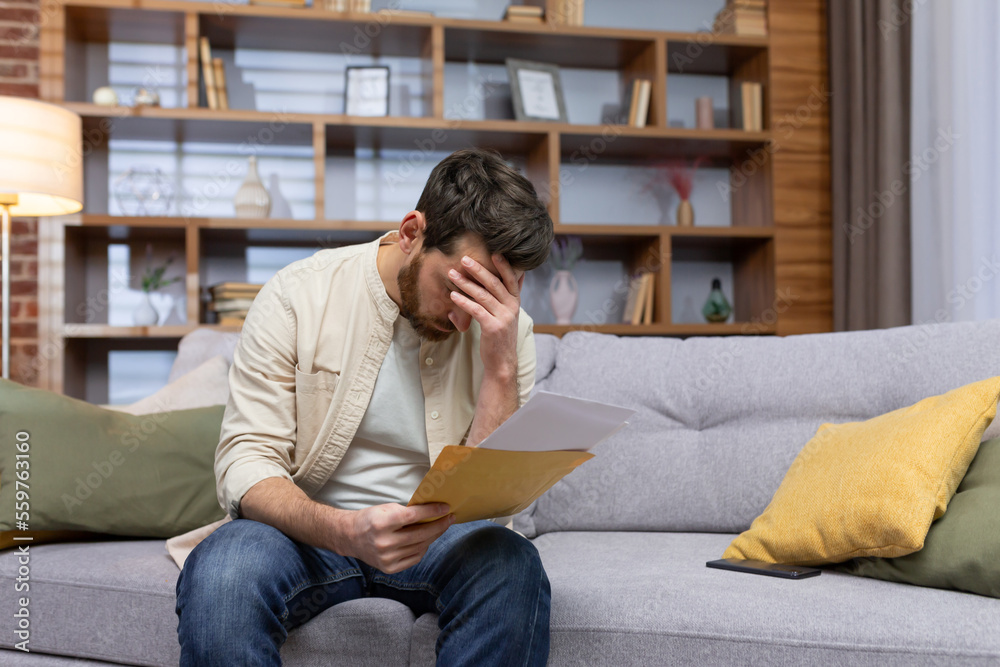Upset man at home sitting on sofa reading letter with bad news notification in envelope opening, unhappy in living room and sad. Upset man at home sitting on sofa reading letter with bad news notification in envelope opening, unhappy in living room and sad.