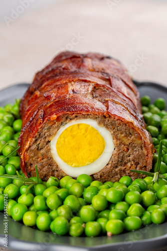 Close-up of baked meatloaf made of ground meat, onion, carrot, stuffed with hard-boiled eggs, with bacon net, served with green peas. German, Scandinavian and Belgian dish. Festive and Easter food. photo