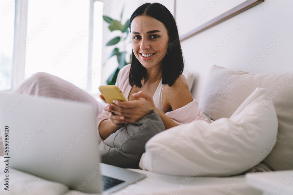 Smiling woman using cellphone and computer while relaxing on bed Smiling woman using cellphone and computer while relaxing on bed