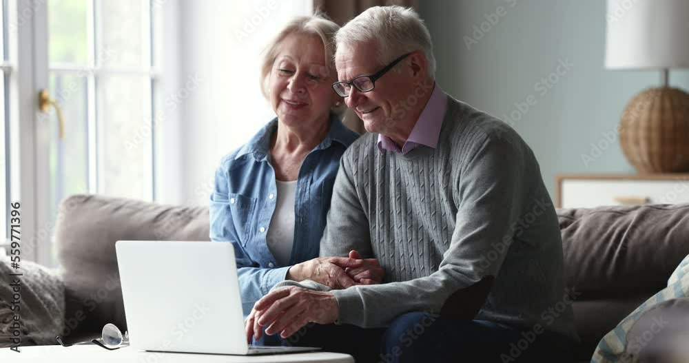 Smiling senior spouses sit on sofa with laptop, buying on-line goods and gifts on internet, discussing news, enjoy modern tech usage, comfort easy electronic services for olders on leisure at home Smiling senior spouses sit on sofa with laptop, buying on-line goods and gifts on internet, discussing news, enjoy modern tech usage, comfort easy electronic services for olders on leisure at home