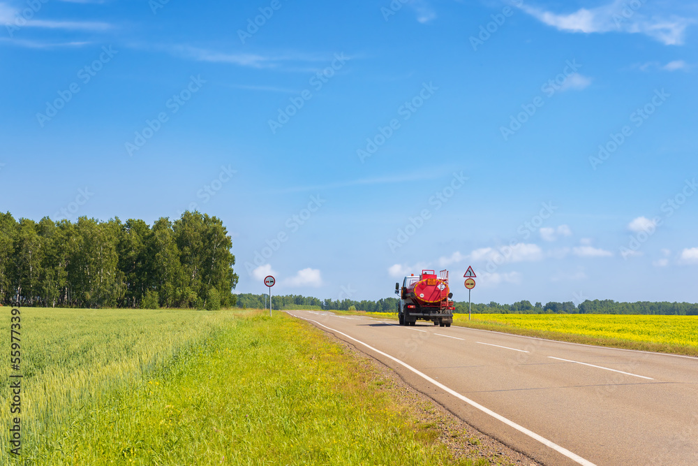 Red fuel truck on an empty rural road among golden flowering farm fields with rapeseed. Summer landscape. Fuel truck is driving along a road. Lorry with orange tanker. Back view.