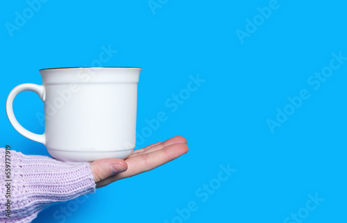 A hand holds a white cup with a hot drink on a blue background. The hand of a woman dressed in a pink sweater holds a ceramic cup on a bright blue background. Free space for text and ads