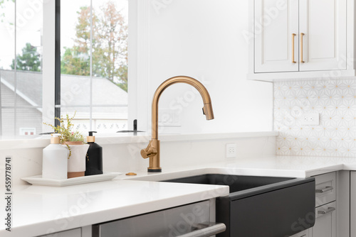 Fotografia A beautiful sink in a remodeled modern farmhouse kitchen with a gold faucet, black apron or farmhouse sink, white granite, and a tiled backsplash