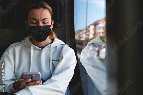 A yong woman in a protective medical mask sits near the window on the bus. Social distance and protection against covid-19 virus in public transport.