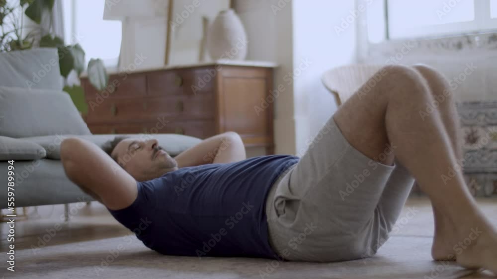 Handsome bearded man doing sit-ups on floor at home. Medium shot of concentrated fit man in T-shirt and shorts taking care of his health, training in morning, doing abs workout. Sport, fitness concept Handsome bearded man doing sit-ups on floor at home. Medium shot of concentrated fit man in T-shirt and shorts taking care of his health, training in morning, doing abs workout. Sport, fitness concept