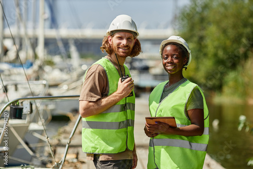 Waist up portrait of two young workers wearing hardhats and smiling at camera while standing in yacht docks